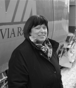 Woman with dark hair, in a winter dress coat, standing next to a VIA Rail traincar.