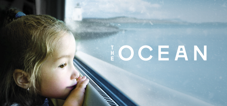 A young girl looking out the window of a train, passing by a coastal scene with snow in the air. Text reads The Ocean.