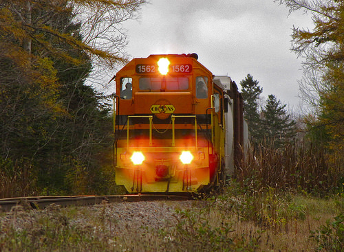 An orange and yellow train with bright headlights heads directly towards the viewer under cloudy fall skies.