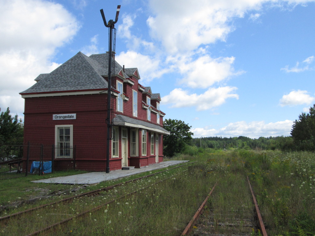 Overgrown by weeds, railway tracks pass the old fashioned red train station at Orangedale Nova Scotia