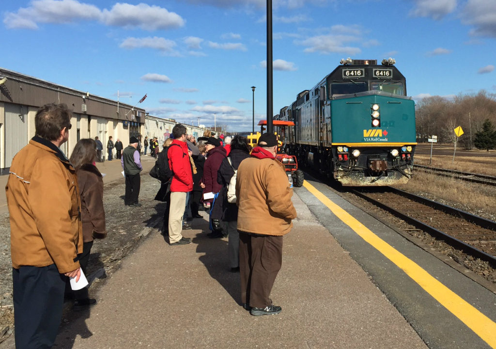 A group of people wait on a station platform as a teal VIA passenger train arrives