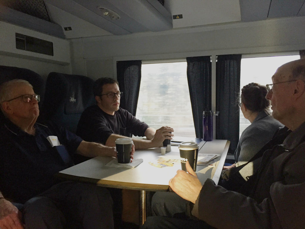 Four people sitting in opposing seats across a table inside a train car, as scenery flies by outside the window